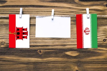 Hanging flags of Gibraltar and Iran attached to rope with clothes pins with copy space on white note paper on wooden background.Diplomatic relations between countries.