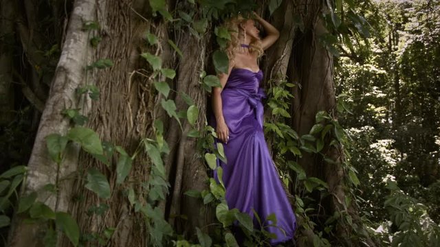 Young romantic woman with curly blonde hair in elegant purple dress posing near big tree and enjoying moment. Dramatic roots and foliage.