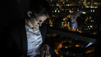 Young man freelancer in glasses is working on his computer near the window in home at night. Self-employed man has overtime job. Freelance and self-employed concept.