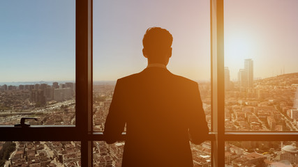 Silhouette of successful young businessman in suit is looking at window. He admires panoramic city...