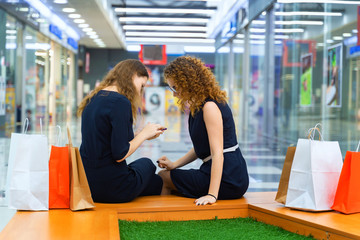 Two young beautiful girls are sitting with packages in a large shopping center and watching the latest information on the phone. Back view