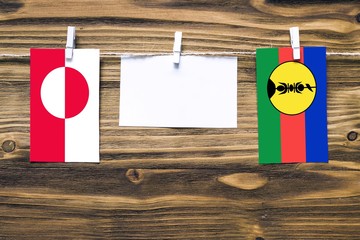 Hanging flags of Greenland and New Caledonia attached to rope with clothes pins with copy space on white note paper on wooden background.Diplomatic relations between countries.