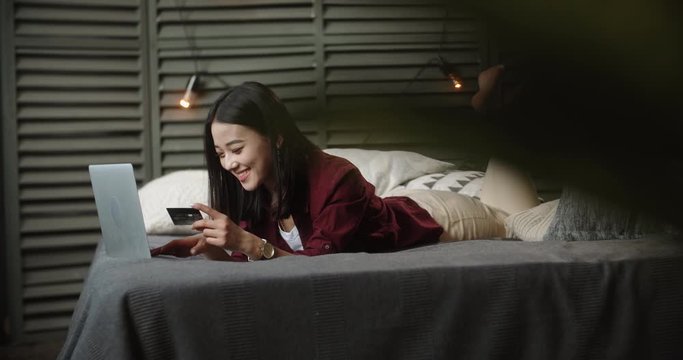Young panasian student spending time on online shopping, lying on large bed with her laptop, keying in credit card and positively smiling - online shopping concept 4k
