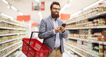 Bearded man with a shopping basket and a mobile phone standing in a supermarket