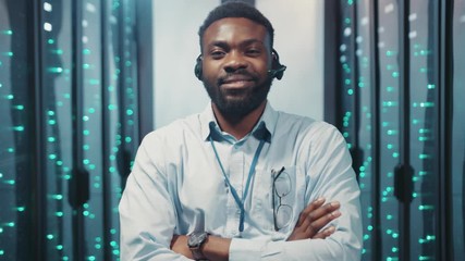 Proud positive african IT engineer crossed arms smiling staying in contemporary computer server room of big data center. - Powered by Adobe