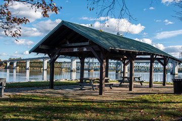 Open shelter in a park with picnic tables along the Kennebec river with a view of a bridge in Bath, Maine on a sunny afternoon with a partly cloudy sky.