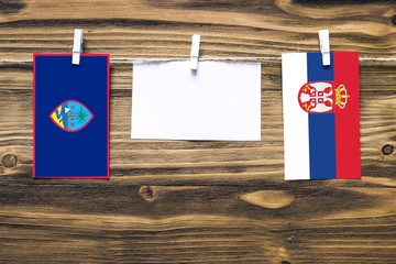 Hanging flags of Guam and Serbia attached to rope with clothes pins with copy space on white note paper on wooden background.Diplomatic relations between countries.