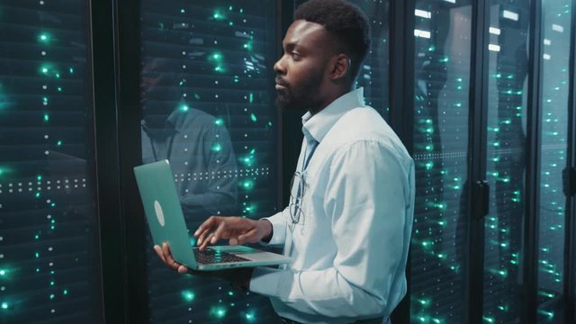 Professional black server engineer running tests on laptop computer inspecting rack server cabinets in big data center storage room.