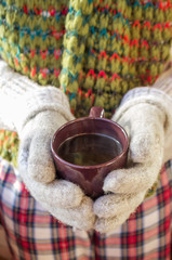 mug with hot drink coffee or tea in the hands of a girl in a sweater and mittens on the background of a frozen winter window, winter mood