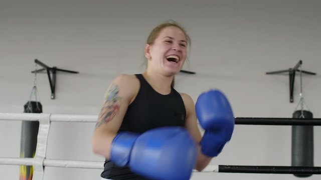 Waist-up arc shot of joyous female boxer in gloves raising hands up and yelling in excitement and then looking at camera with smile and throwing punches in the air on ring