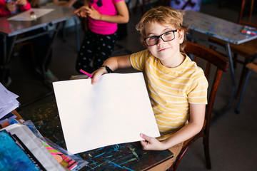 Blonde boy with glasses drawing. Group of elementary school pupils in classroom on art class. Paper...