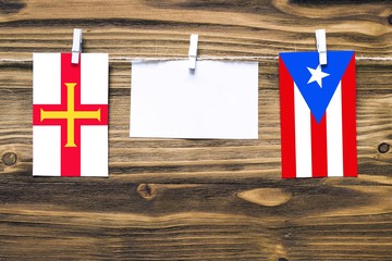 Hanging flags of Guernsey and Puerto Rico attached to rope with clothes pins with copy space on white note paper on wooden background.Diplomatic relations between countries.