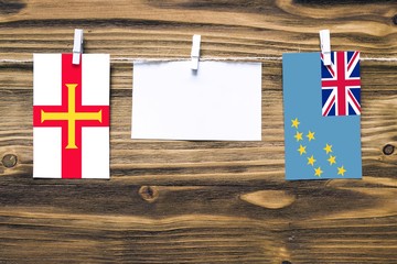 Hanging flags of Guernsey and Tuvalu attached to rope with clothes pins with copy space on white note paper on wooden background.Diplomatic relations between countries.