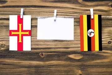 Hanging flags of Guernsey and Uganda attached to rope with clothes pins with copy space on white note paper on wooden background.Diplomatic relations between countries.