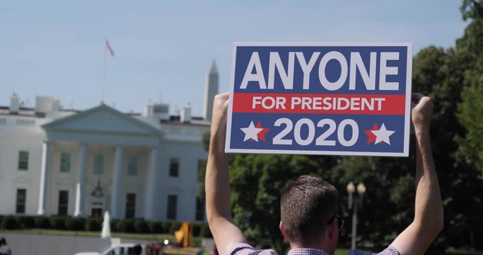 A man holds an Anyone 2020 election protest sign in front of the White House on a sunny summer day.  	
