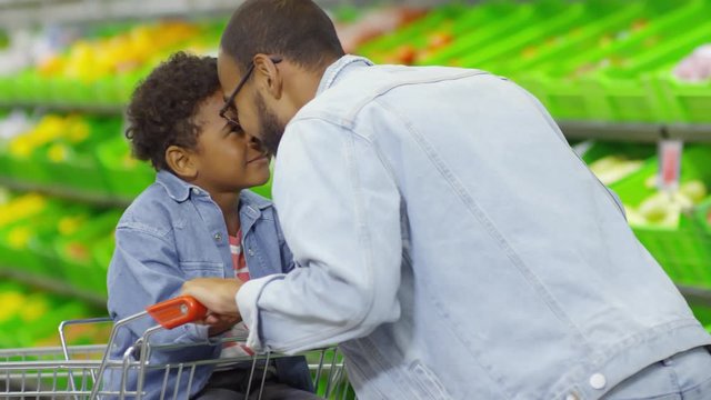 Medium shot of loving father rubbing noses with little son of primary school age while shopping at supermarket, boy sitting in shopping cart