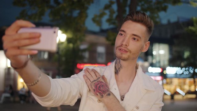 Tattoed young man with stylish hairstyle is taking selfie outdoors with smartphone samera standing alone at night in summer. People and photograph concept.
