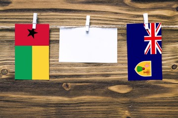 Hanging flags of Guinea Bissau and Turks And Caicos Islands attached to rope with clothes pins with copy space on white note paper on wooden background.Diplomatic relations between countries.