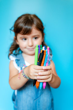 Portrait of cute little girl picking up a handful of colored ballpoint pens on blue background