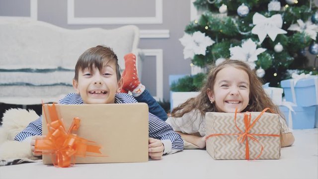 Careless, smiling small siblings enjoying Christmas time, laying on the carpet under fir-tree, showing their heads from behind two giftboxes.