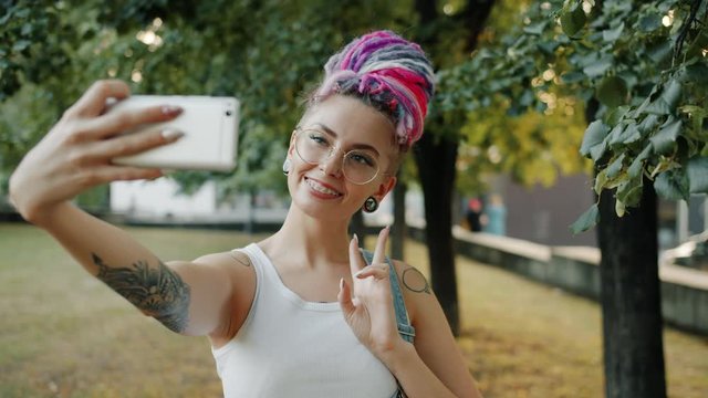 Trendy young lady hipster is taking selfie in city park posing with hand gestures smiling posing for smartphone camera outdoors. People and technology concept.