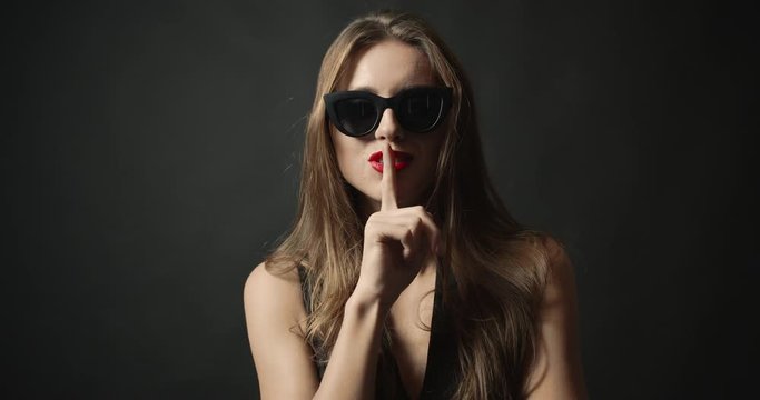 Young seductive woman in sunglasses and dark dress whispering on black background, fashion and glamour
