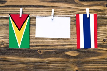 Hanging flags of Guyana and Thailand attached to rope with clothes pins with copy space on white note paper on wooden background.Diplomatic relations between countries.