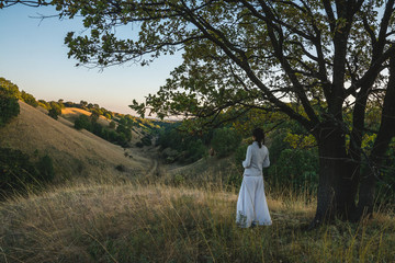 Young woman in white clothing standing under the tree and watching autumn evening sky over the hills of Deliblatska pescara (Deliblato sands) in Serbia