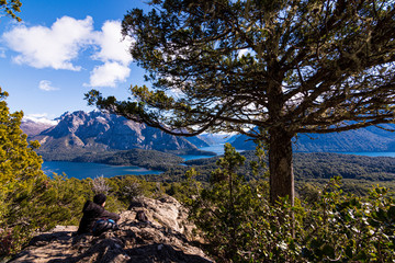 Young couple resting on the rocks beneath the shadow of a coihue tree against Andes range and Nahuel Huapi lake in Bariloche, Patagonia, Argentina 