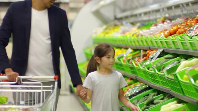 Little Asian girl holding father&rsquo;s hand, taking squash from shelf at supermarket and putting it into shopping cart