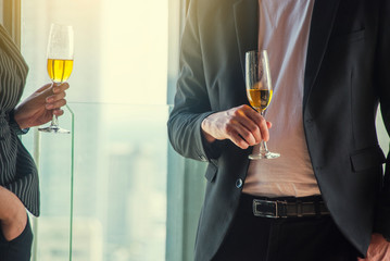 Colleagues, business men, wearing suits, talking to business women, both holding white glasses, standing at the party.