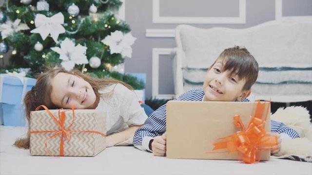Careless, smiling small siblings enjoying Christmas time and presents, laying on the carpet under fir-tree.