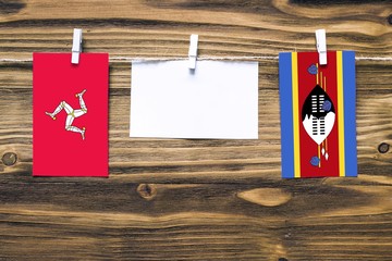 Hanging flags of Isle Of Man and Swaziland attached to rope with clothes pins with copy space on white note paper on wooden background.Diplomatic relations between countries.