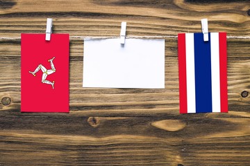 Hanging flags of Isle Of Man and Thailand attached to rope with clothes pins with copy space on white note paper on wooden background.Diplomatic relations between countries.