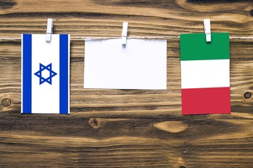 Hanging flags of Israel and Italy attached to rope with clothes pins with copy space on white note paper on wooden background.Diplomatic relations between countries.