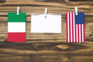 Hanging flags of Italy and United States attached to rope with clothes pins with copy space on white note paper on wooden background.Diplomatic relations between countries.