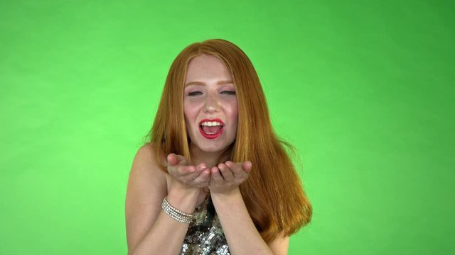 In front of a green screen in studio, a young women blows a handful of red metallic confetti towards the camera