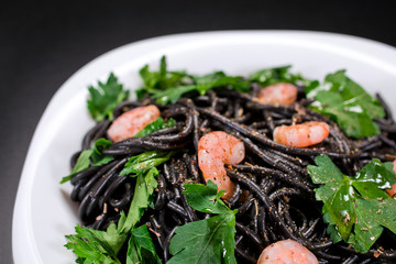 Black long spaghetti on a plate with shrimps, green parsley and spices on black background.