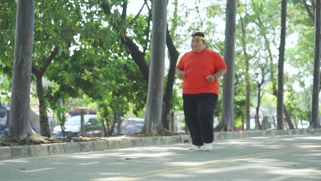 JAKARTA, Indonesia - October 28, 2019: Obese man running at park and taking a rest by sitting on the road under green trees. Shot in 4k resolution