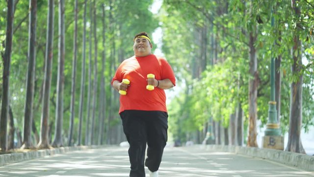 Overweight young man running at the park while carrying two dumbbells and wearing sportswear. Shot in 4k resolution