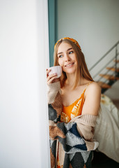 Beautiful smiling young woman fair long hair girl wearing in cozy knitted cardigan with cup of tea near window at home