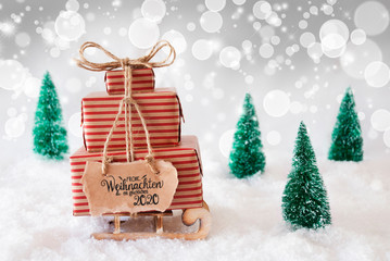 Sled With Christmas Gift And Label With German Frohe Weihnachten Und Ein Glueckliches 2020 Means Merry Christmas And A Happy 2020. Snow With Christmas Tree And Gray Sparkling Background