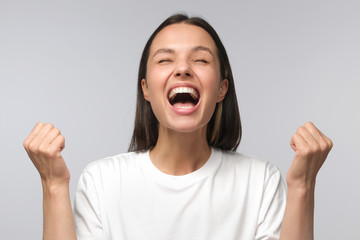 Close-up of emotional european woman showing white teeth while screaming with joy, holding hands in gesture of winner, isolated on gray background