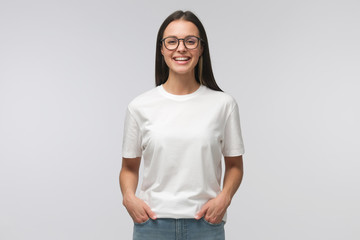 Young laughing woman standing with hands in pockets, wearing blank white t-shirt with copy space,...