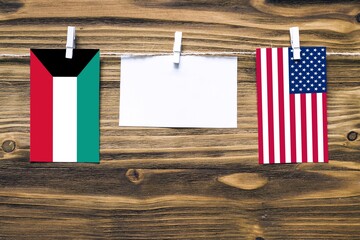 Hanging flags of Kuwait and United States attached to rope with clothes pins with copy space on white note paper on wooden background.Diplomatic relations between countries.