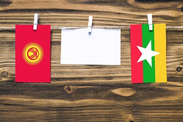 Hanging flags of Kyrgyzstan and Myanmar attached to rope with clothes pins with copy space on white note paper on wooden background.Diplomatic relations between countries.