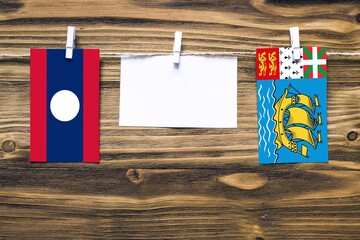 Hanging flags of Laos and Saint Pierre And Miquelon attached to rope with clothes pins with copy space on white note paper on wooden background.Diplomatic relations between countries.