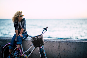 Happy beautiful curly healthy caucasian adult middle age woman sit down on a vintage coloured bike and enjoy the ocean in outdoor leisure activity - trendy people with hapy life - obrazy, fototapety, plakaty