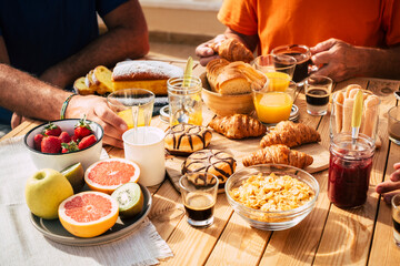 Close up of group of unrecognizable people eating brekfast all together on a wooden table under the sun -outdoor food activity and vacation family concept - fruit coffee and brioches