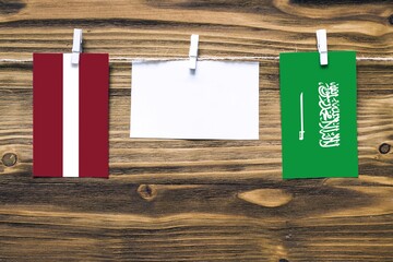 Hanging flags of Latvia and Saudi Arabia attached to rope with clothes pins with copy space on white note paper on wooden background.Diplomatic relations between countries.
